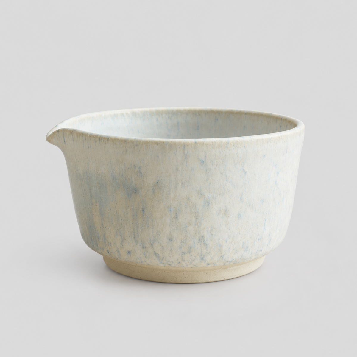 A ceramic matcha bowl with a small pouring spout, featuring a textured, speckled glaze in light blue and cream tones, set against a plain light gray background.