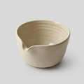 A handmade ceramic matcha bowl with a small pouring spout, featuring a textured, ribbed interior and a matte, earthy beige finish, set against a solid light gray background.