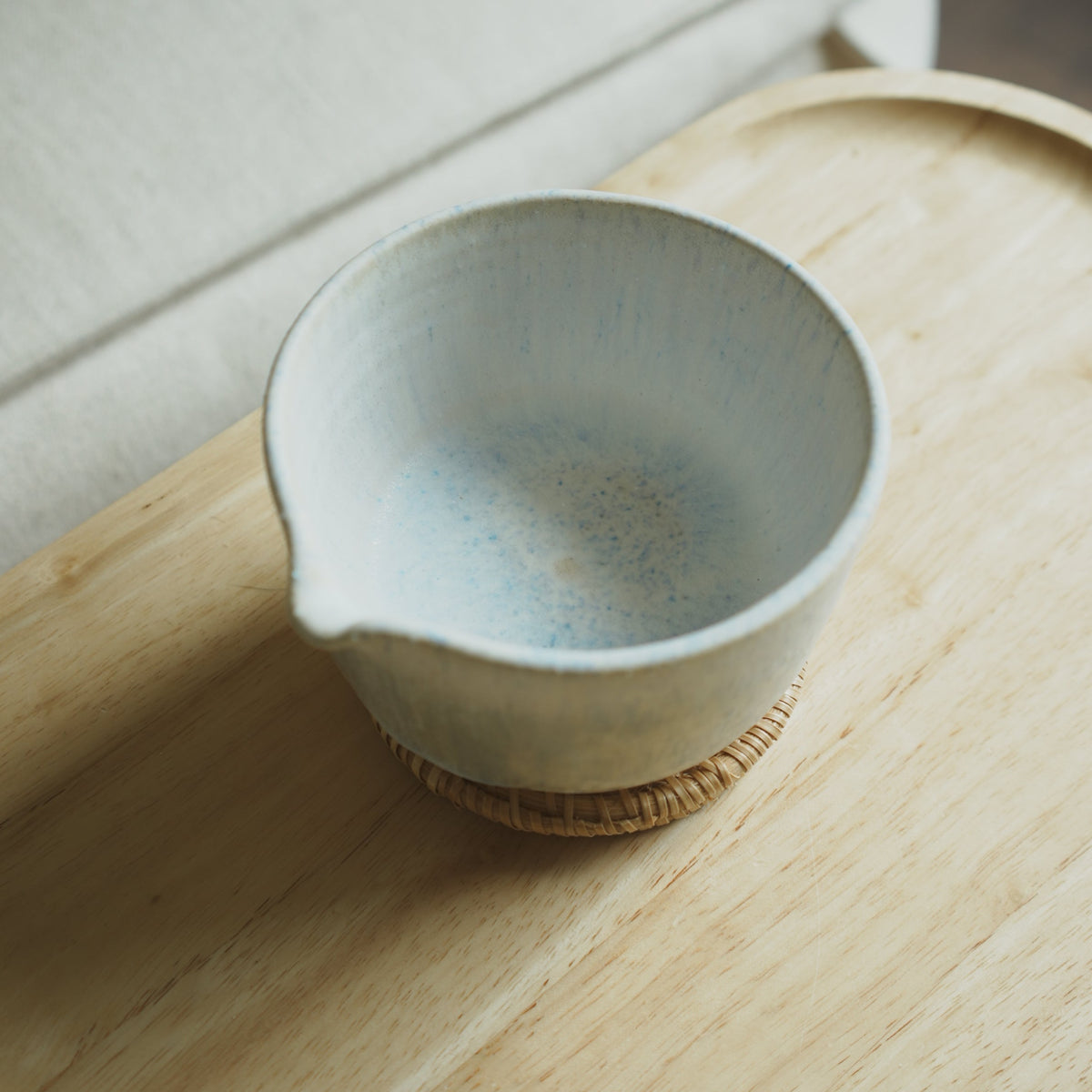A handmade ceramic matcha bowl with a light blue-gray glaze and a small spout, resting on a woven coaster inside a light wood tray.