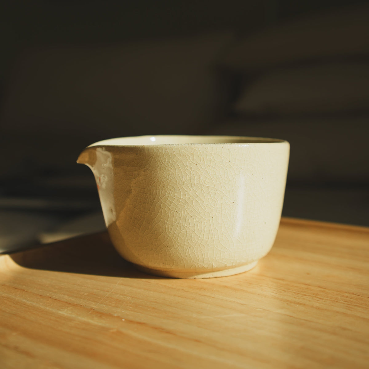 A small beige ceramic matcha bowl with a spout, resting on a wooden table in warm, directional sunlight.