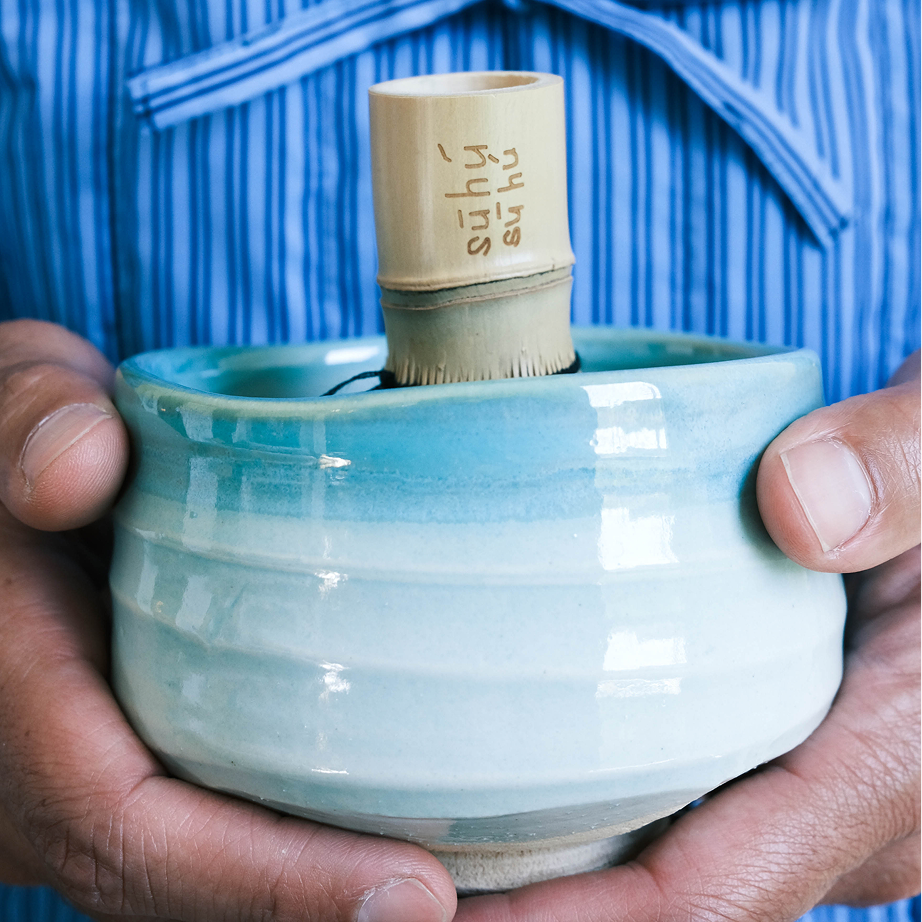 Person holding a light blue ceramic bowl with a bamboo matcha whisk against a blue striped jacket.