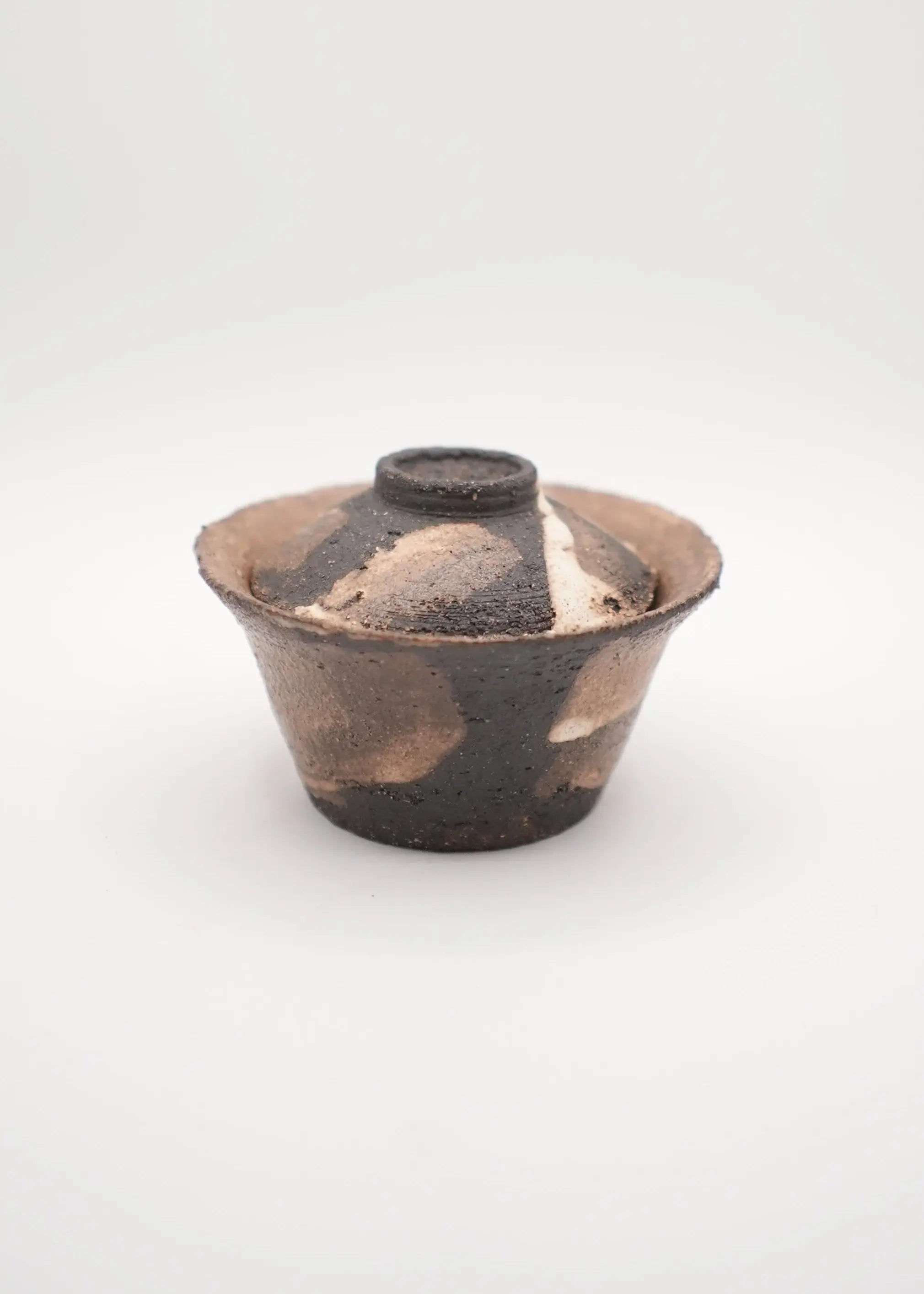 A handmade ceramic lidded bowl with a rustic, hand-painted finish in dark brown and earthy beige tones, set against a plain light gray background.
