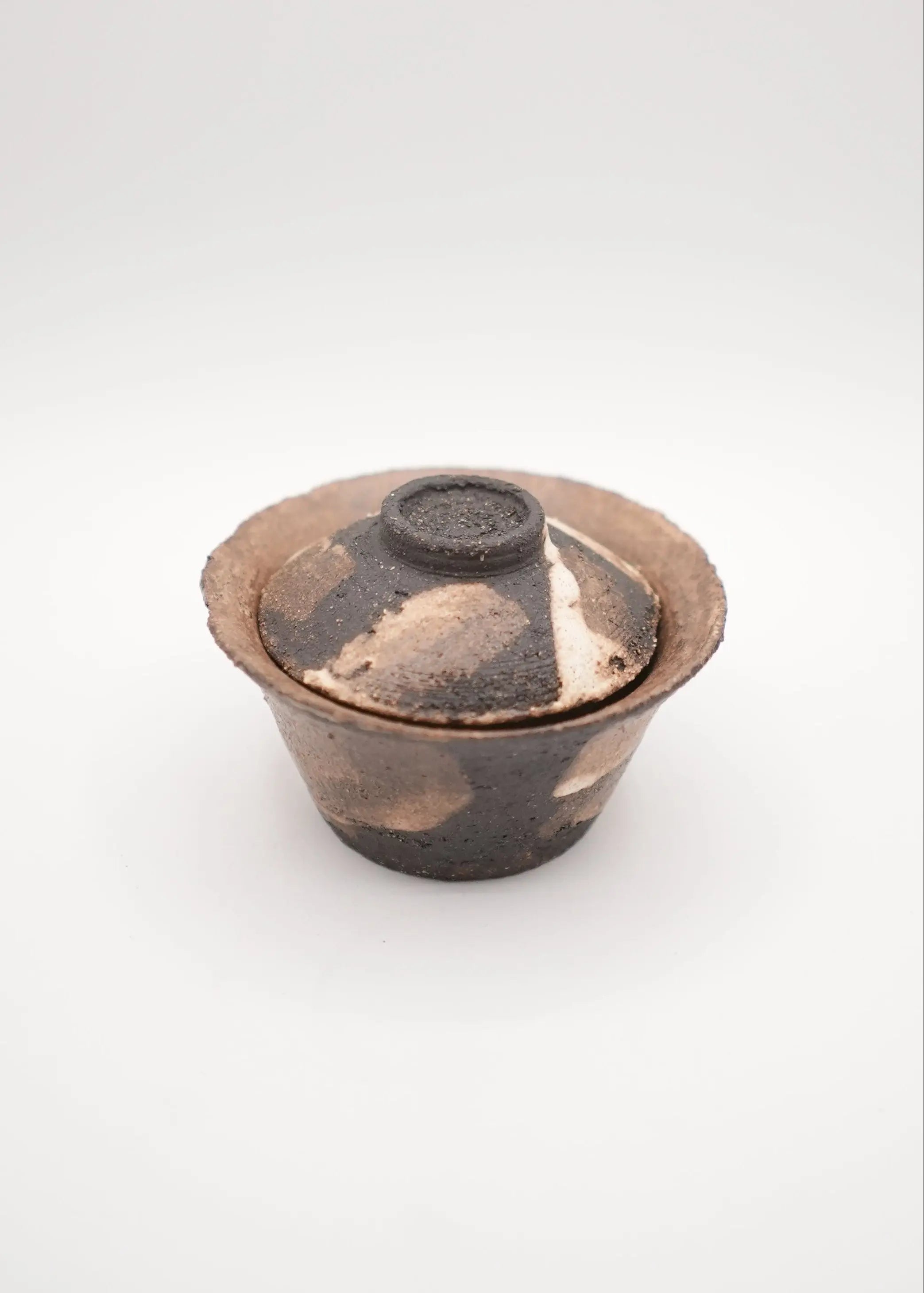A small handmade ceramic lidded cup or bowl with a textured, rustic finish in earthy brown, black, and beige tones, set against a plain light gray background.