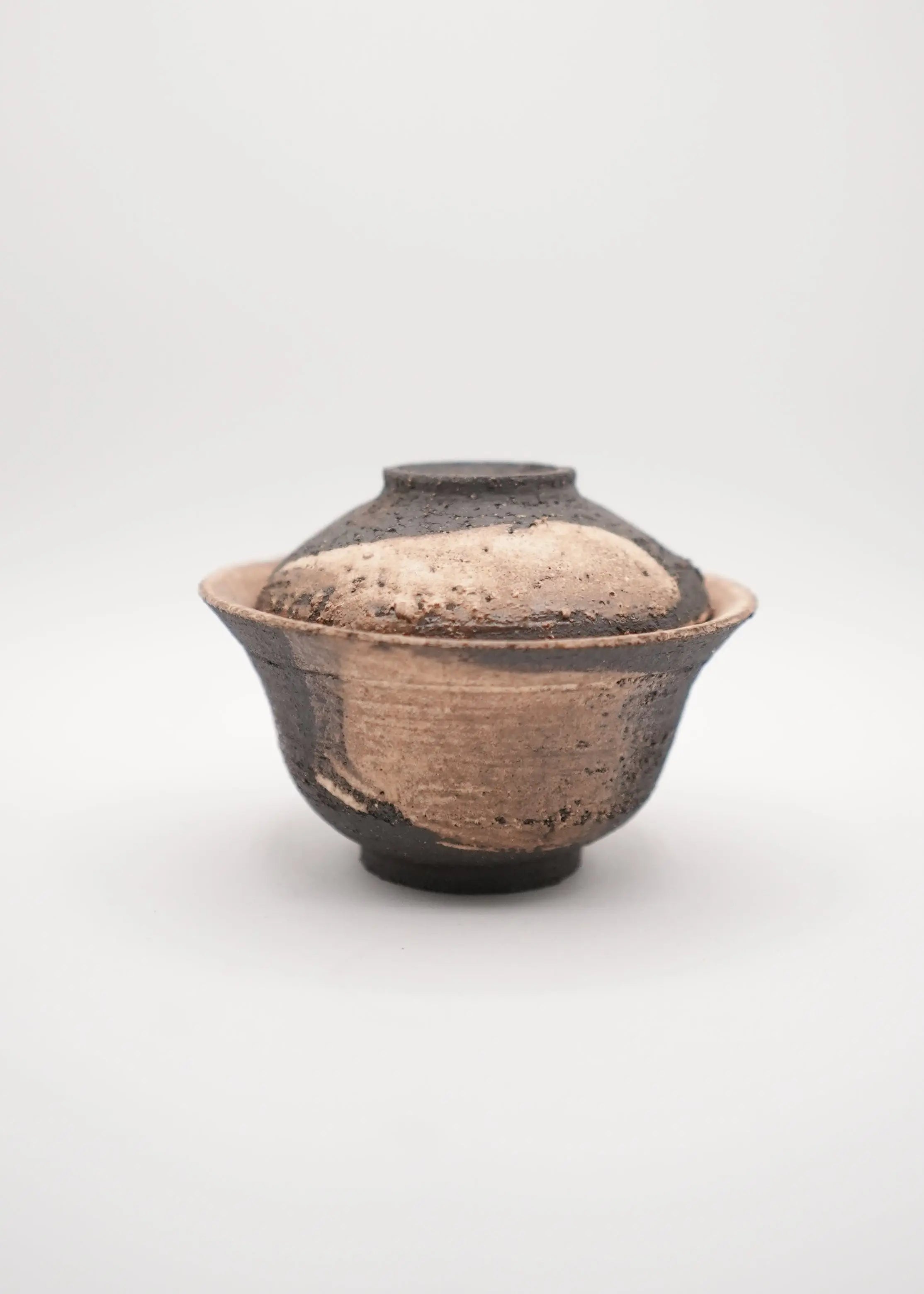 A handmade ceramic lidded bowl with a rustic, textured finish in earthy brown and black tones, photographed against a seamless light gray background.