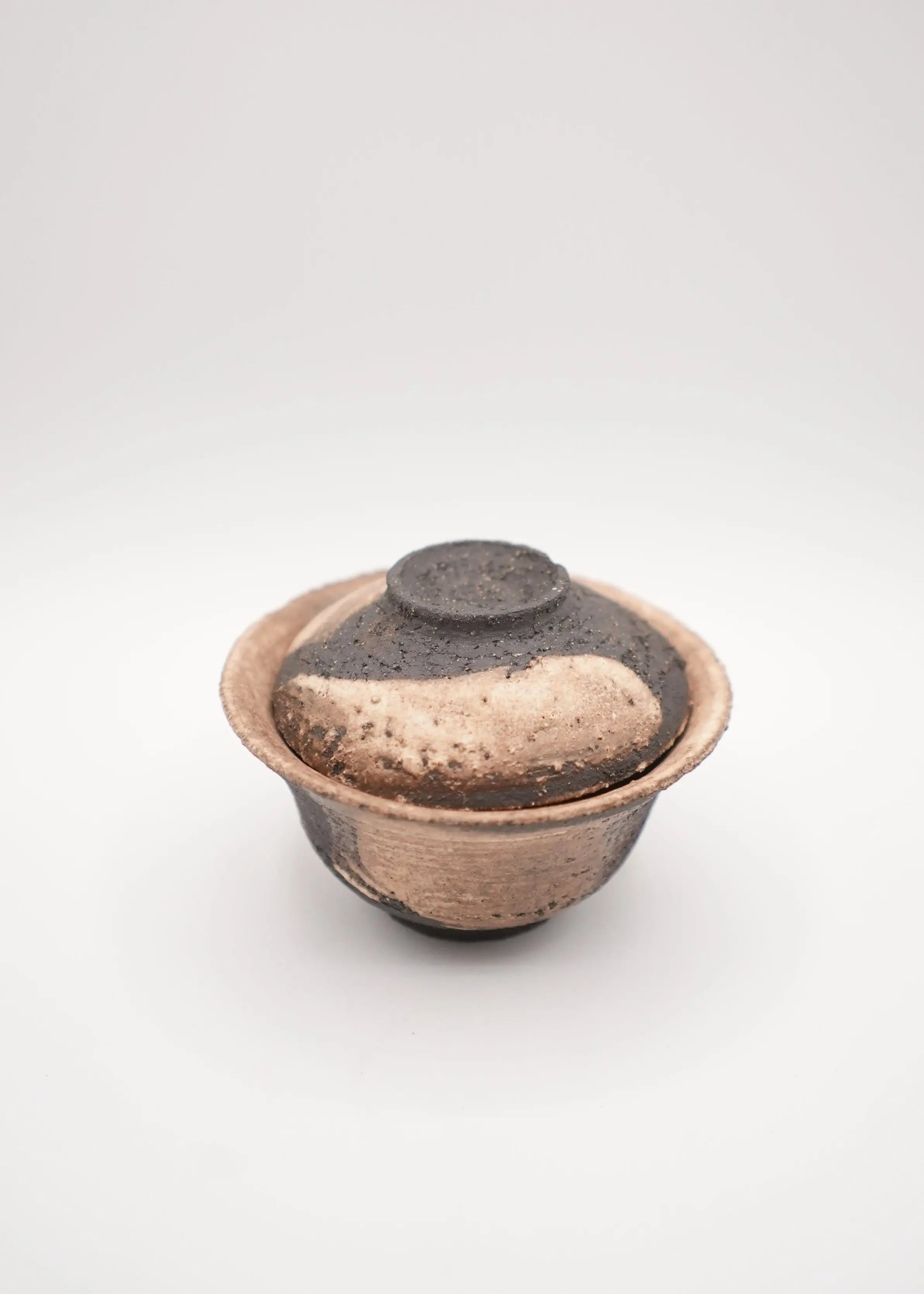 A small handmade ceramic lidded bowl with a textured, speckled finish in earthy brown, beige, and dark charcoal tones, photographed on a seamless light gray background.