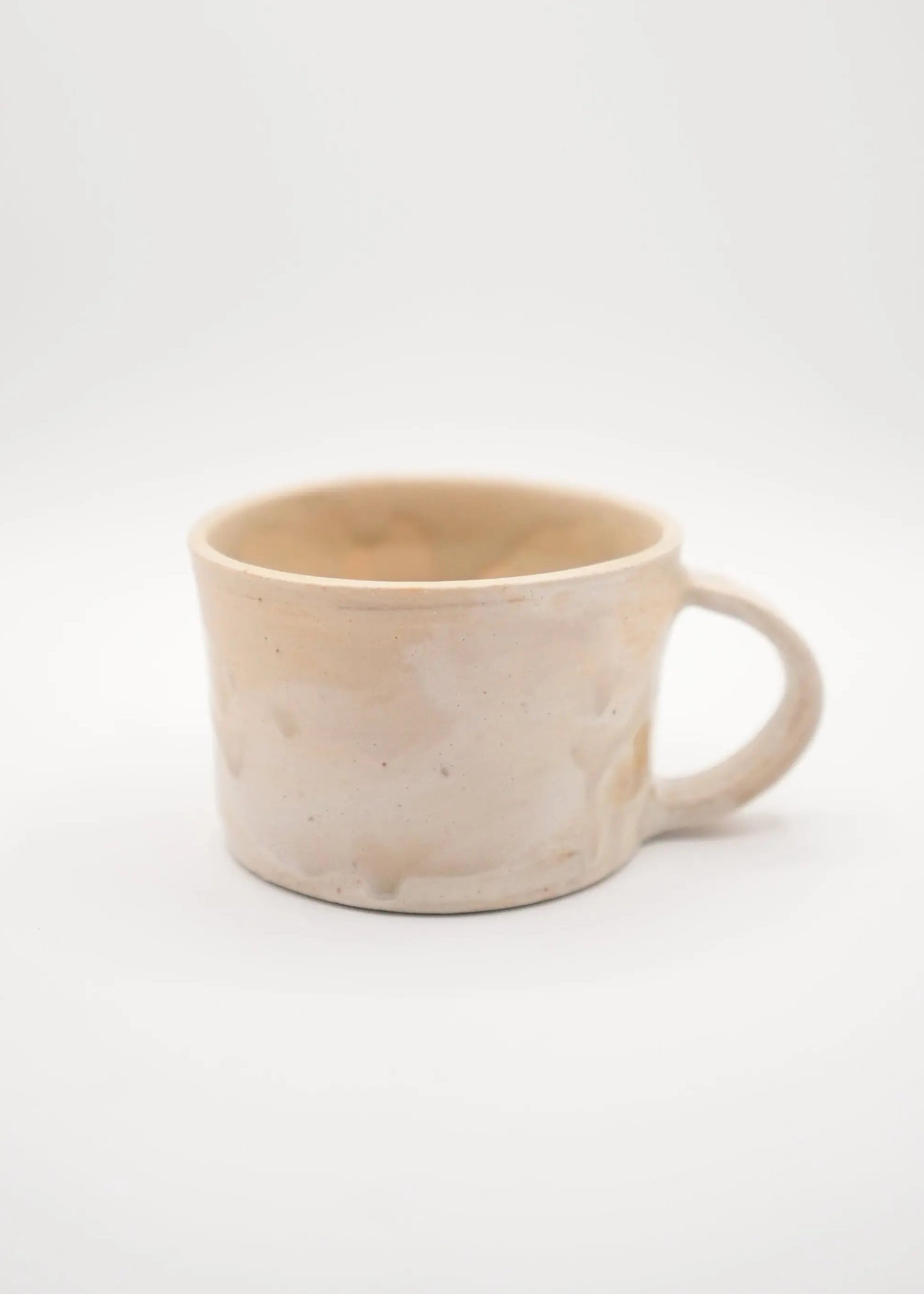 A handmade ceramic mug with a textured, speckled beige and cream glaze, featuring a simple C-shaped handle, set against a seamless light gray background.