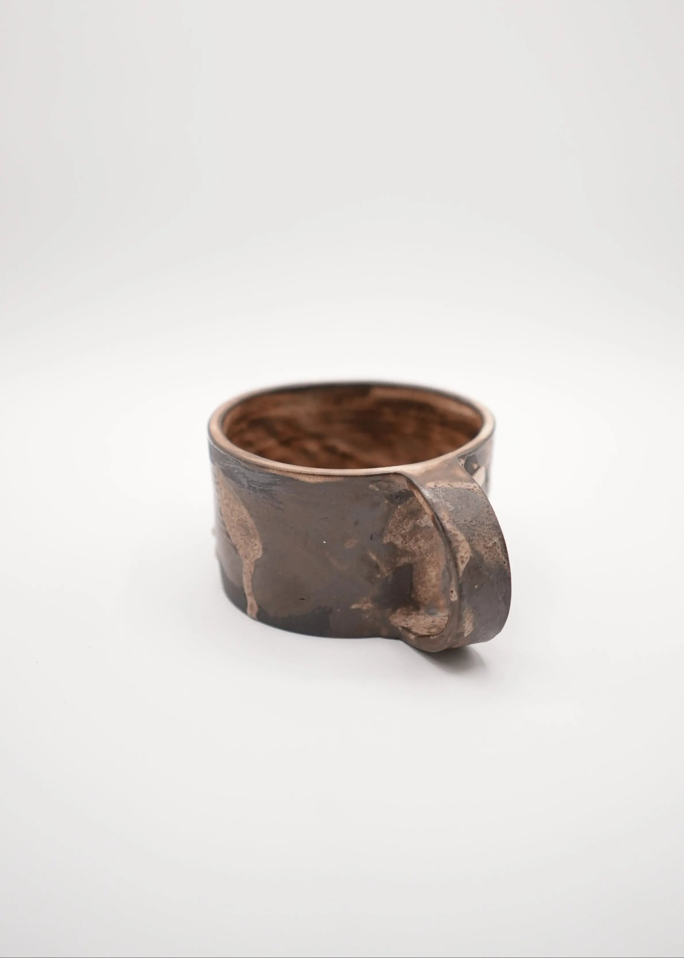 A rustic handmade ceramic mug with a dark brown and black textured glaze, featuring a small integrated handle on the right side, photographed on a seamless light gray background.