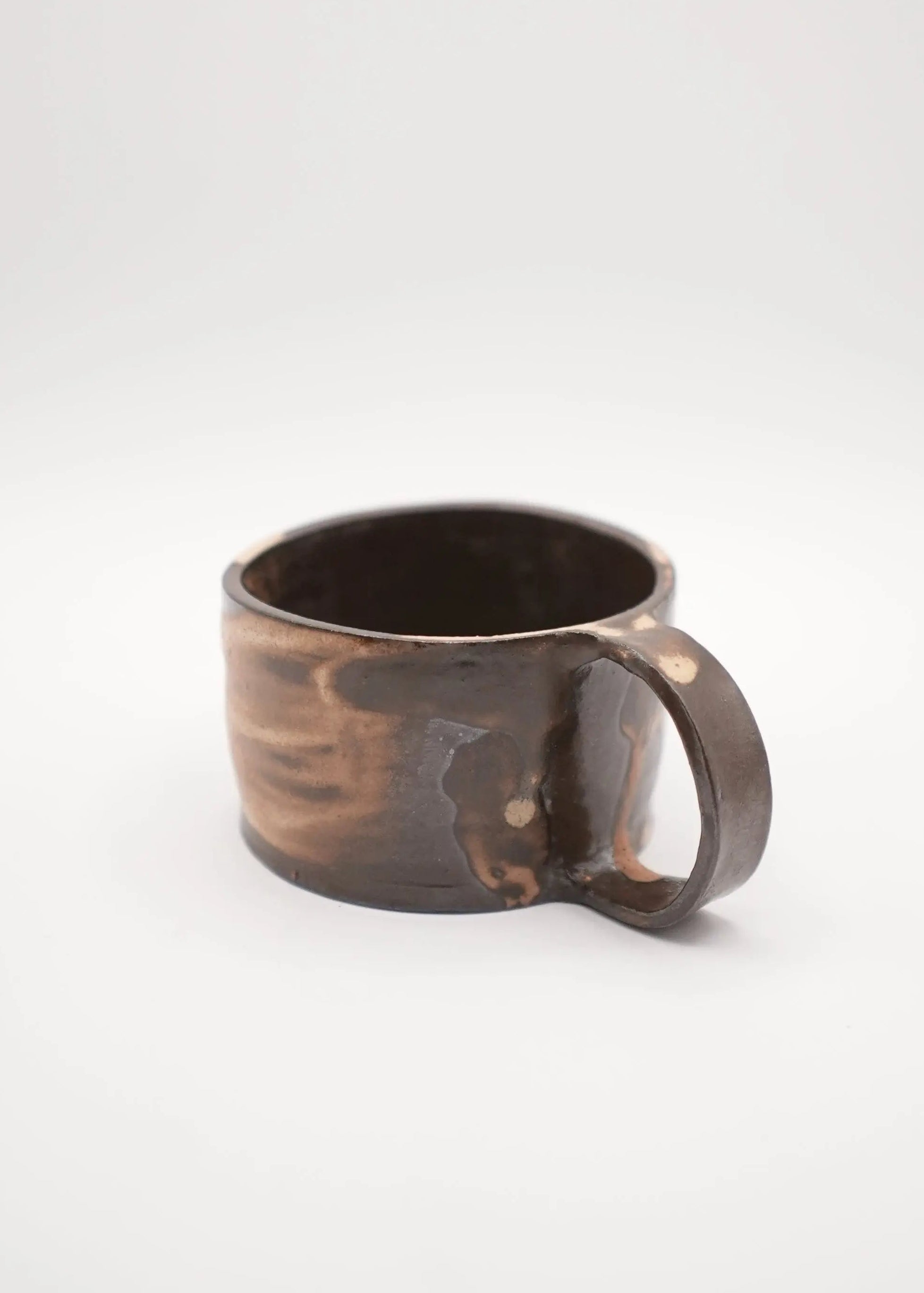 A handmade ceramic mug with a rustic, hand-painted dark brown and black finish, featuring a thick integrated handle, shown on a seamless light gray background.