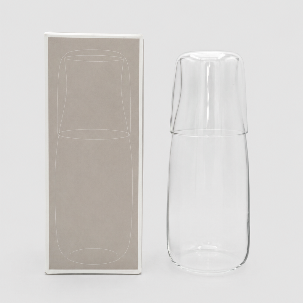 Clear glass carafe and cup set next to packaging gift box on a light grey background