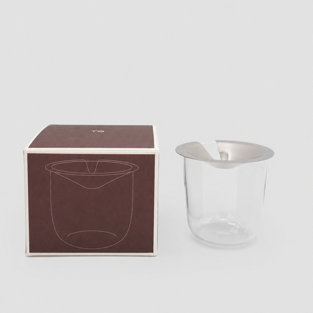 Clear glass vase with stainless steel lid next to brown packaging box on a light grey background